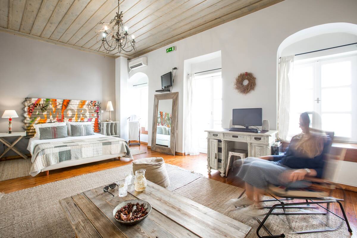 A spacious room with a bed, a desk, and a television. There is a chandelier, a mirror, and a wreath on the wall.