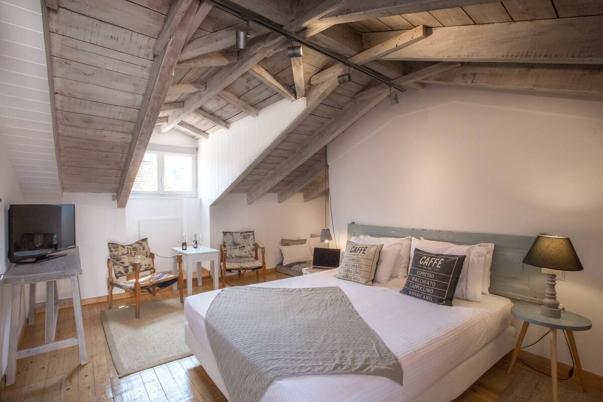 A cozy bedroom with a large bed, a desk, a chair, a TV, and a lamp.