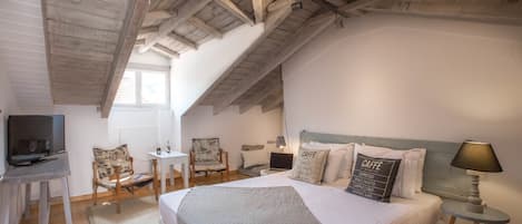 A cozy bedroom with a large bed, a desk, a chair, a TV, and a lamp.