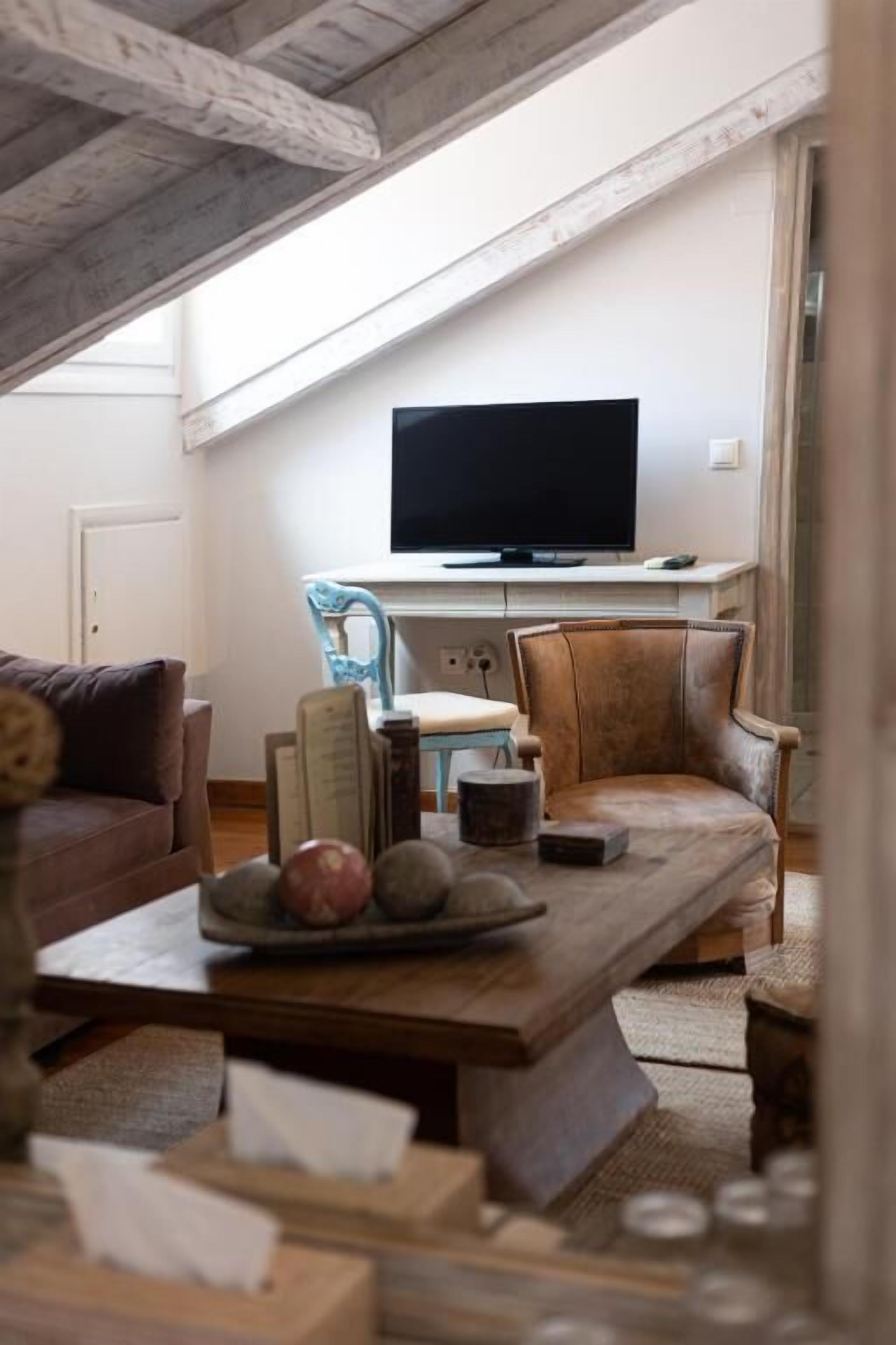 A cozy living room with a wooden ceiling, a flat-screen TV on a white stand, a brown leather armchair, a blue chair, and a wooden coffee table with decorative items.