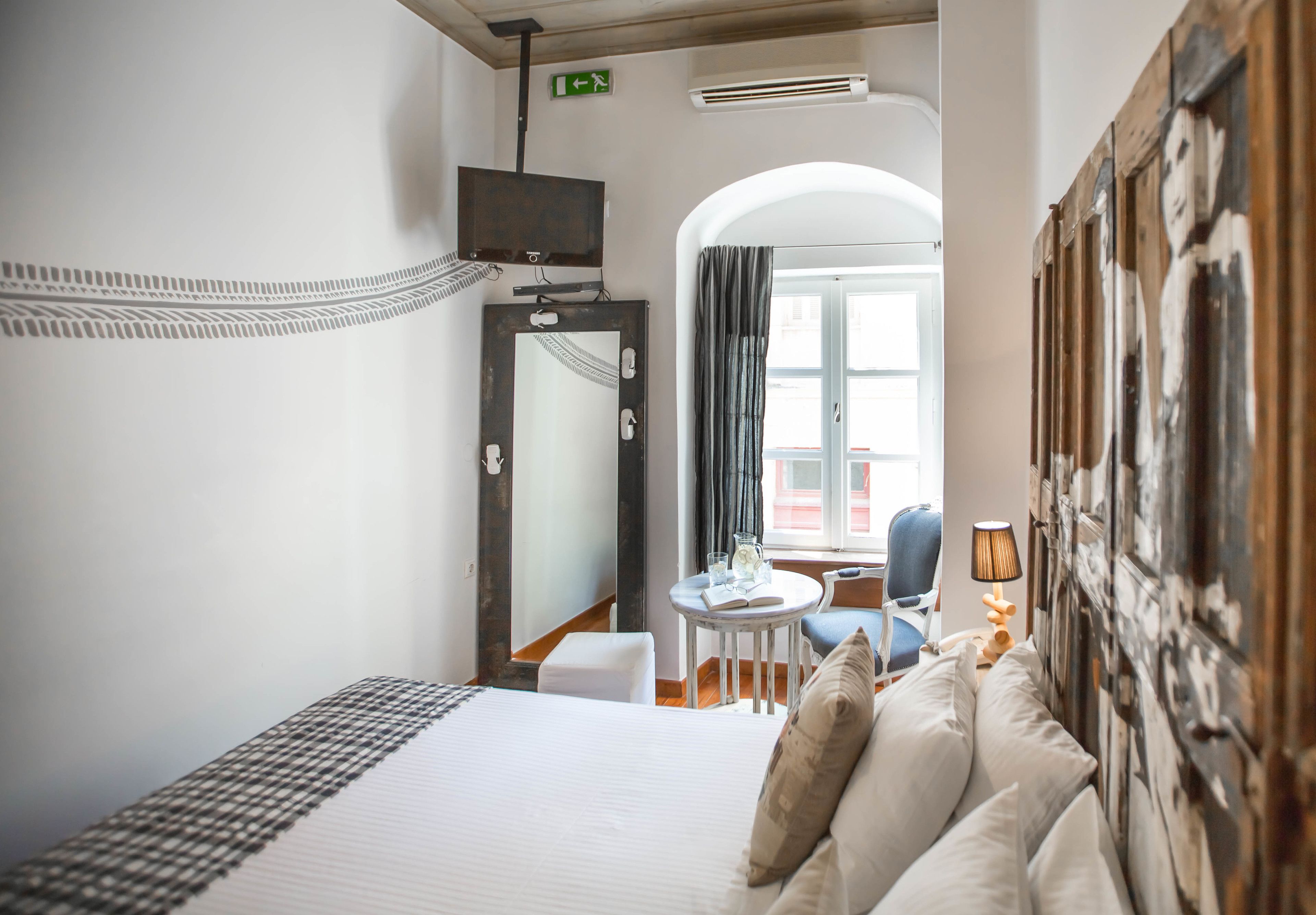 A hotel room with a bed, a chair, a small table with a lamp, a mirror, and a view of the outside.