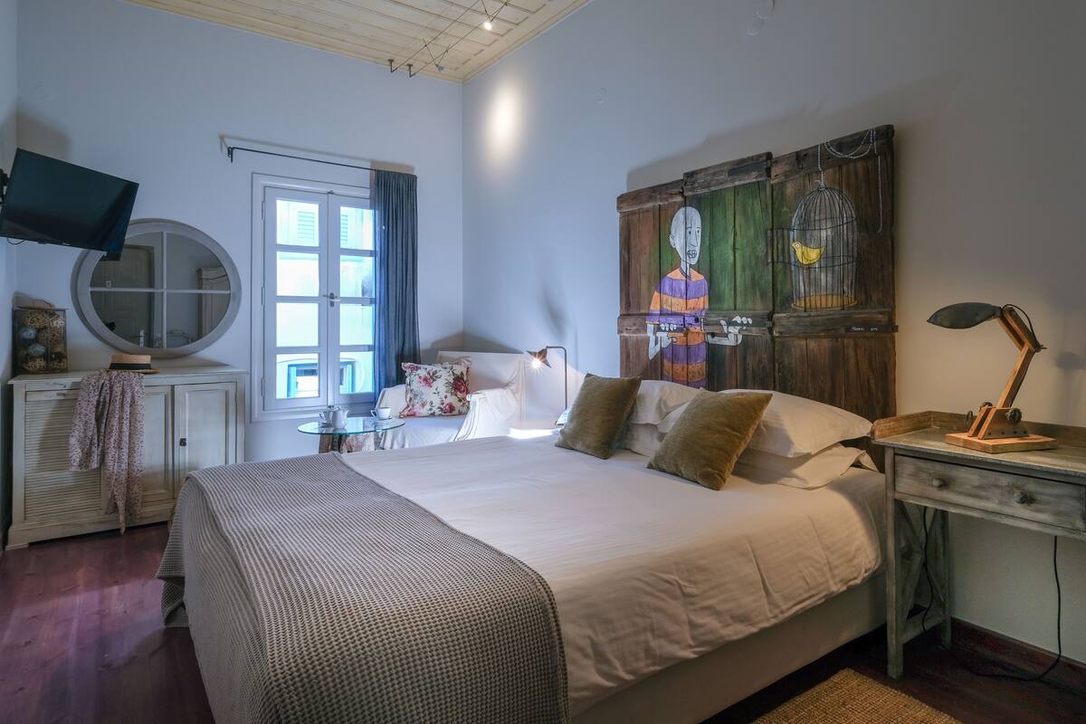 A bedroom with a large bed, a round mirror, a television, a window with curtains, and a wooden artwork on the wall.