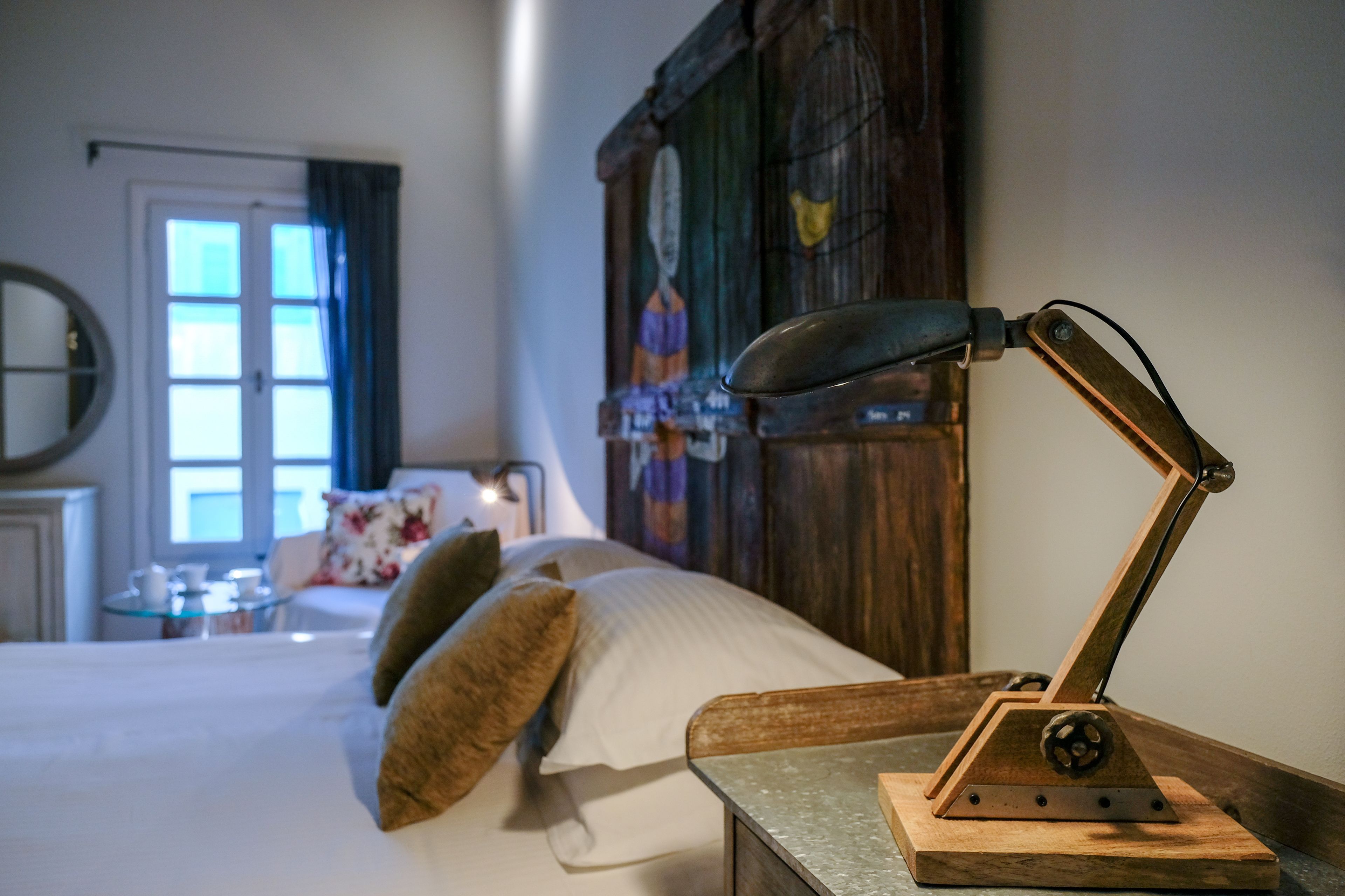 A neatly made bed with pillows, a wooden nightstand with a lamp, a wooden headboard, a window with curtains, and a small table with cups and a book.