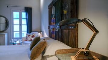 A neatly made bed with pillows, a wooden nightstand with a lamp, a wooden headboard, a window with curtains, and a small table with cups and a book.