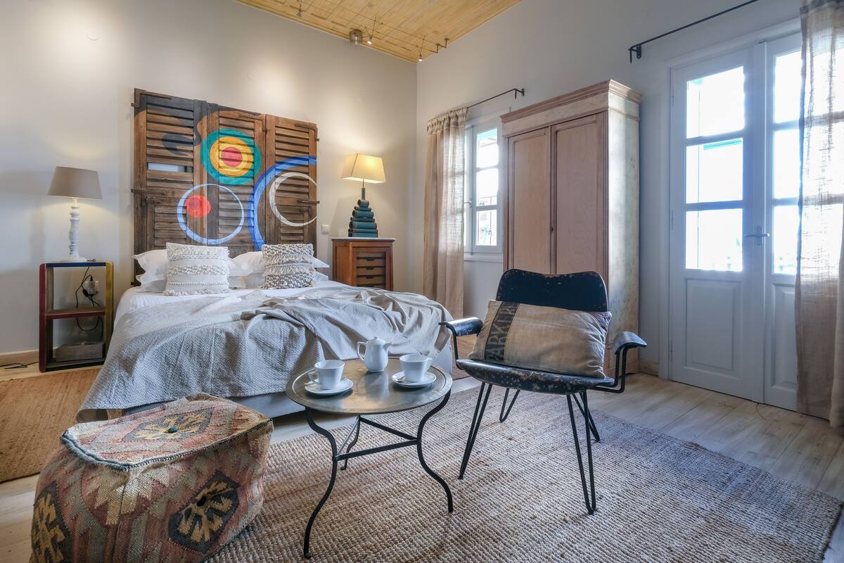 A bedroom with a bed, bedside tables, a lamp, a chair, a small table with cups, and a window with curtains.