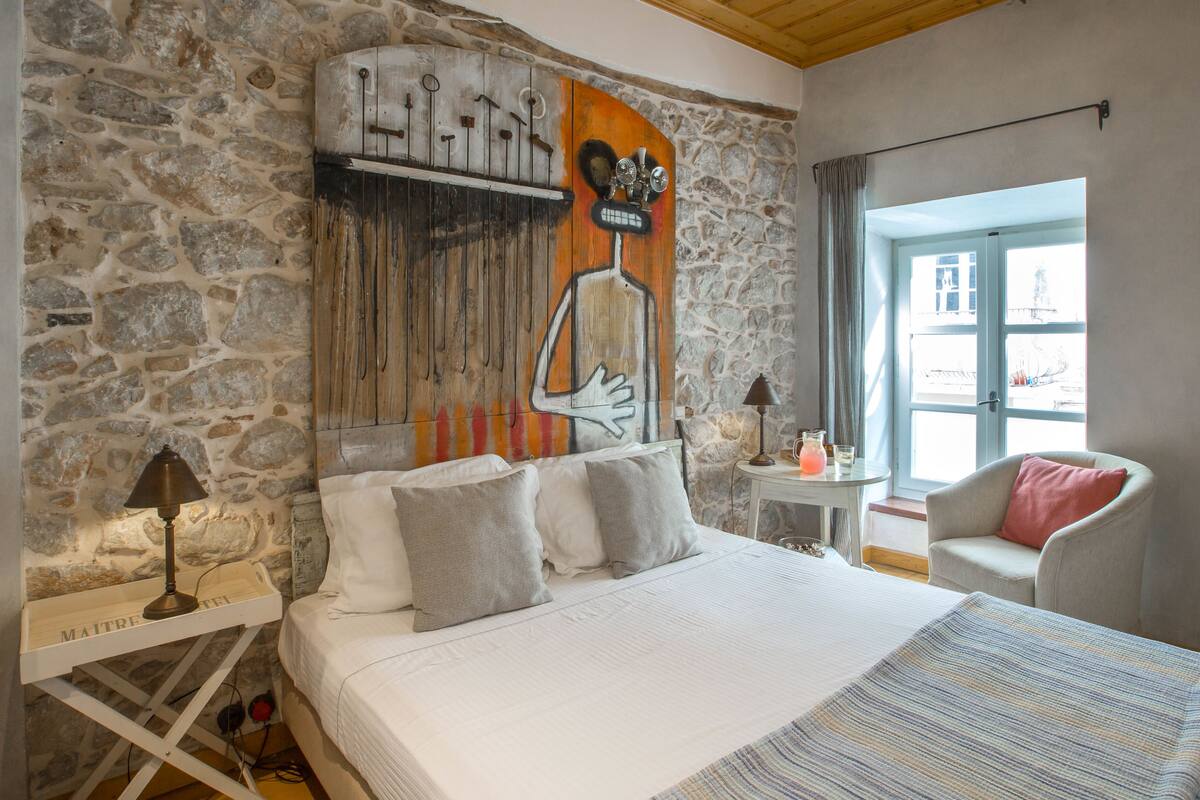 A bedroom with a stone wall, a bed with pillows, a nightstand with a lamp, a chair, and a window with a view.