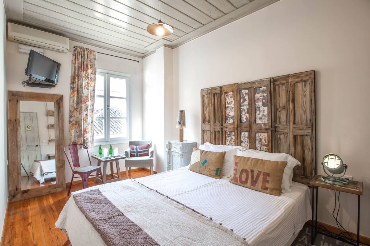 A bedroom with a bed, a wooden headboard, a desk with a chair, a television, and a window with curtains.