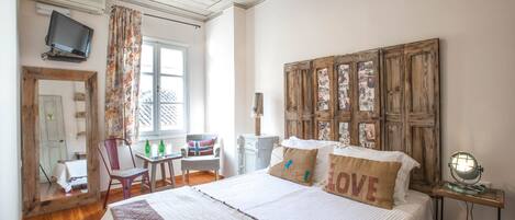 A bedroom with a bed, a wooden headboard, a desk with a chair, a television, and a window with curtains.