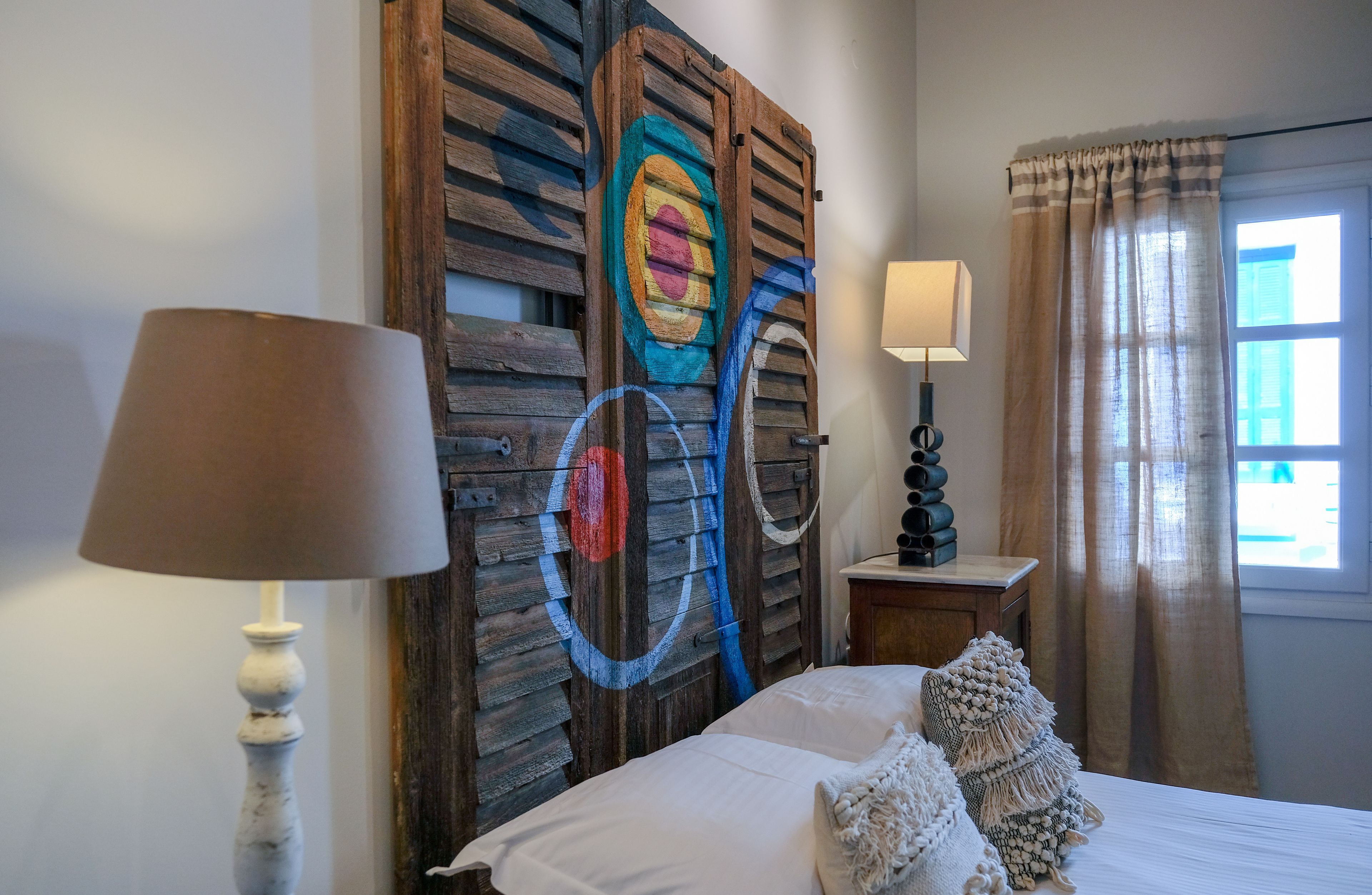A bedroom with a wooden headboard, a bedside table with a lamp, a window with curtains, and a bed with pillows.