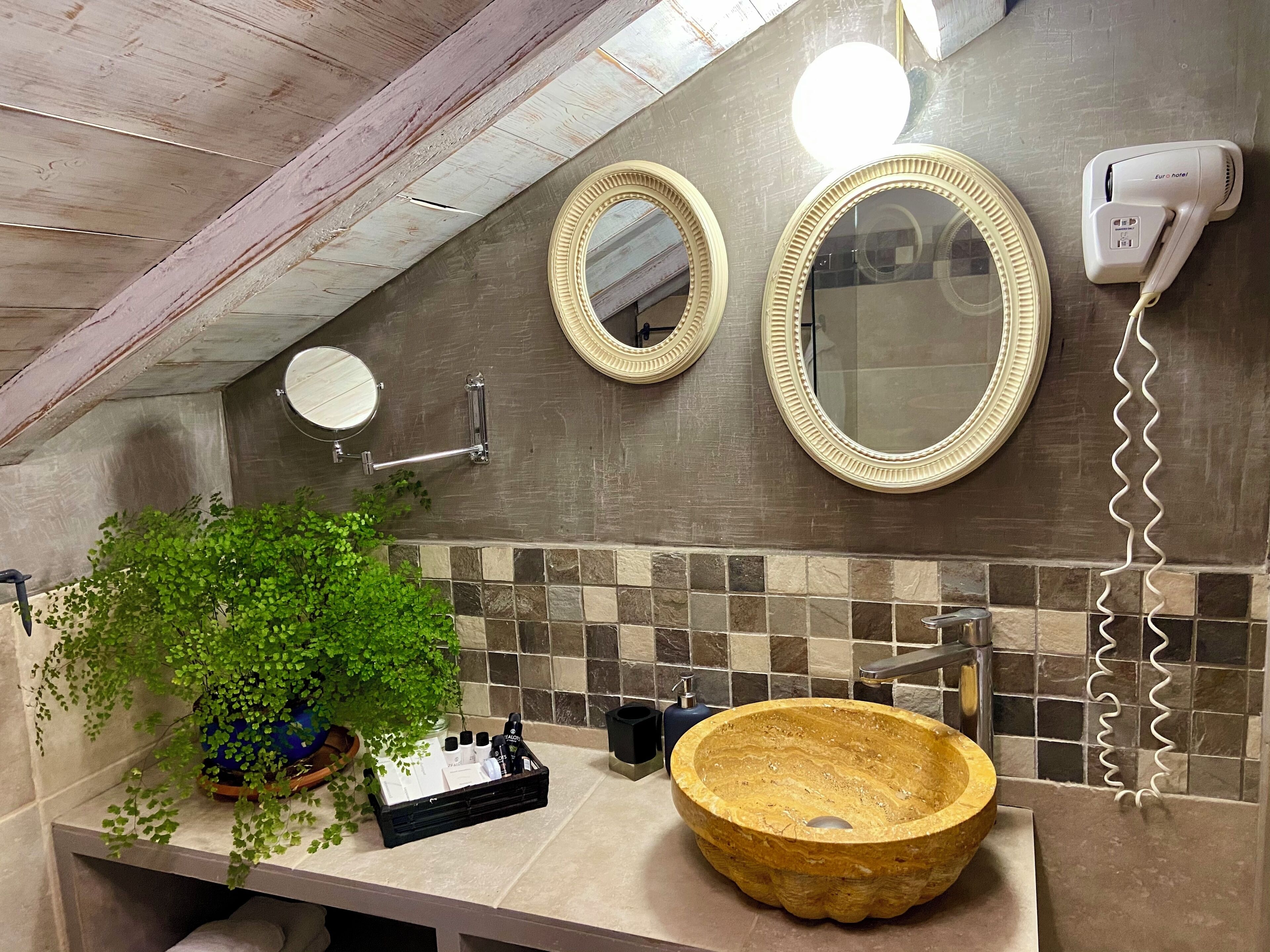 Baño con techo de madera, un espejo grande, secador de pelo, lavabo con lavamanos de piedra y una planta en maceta.