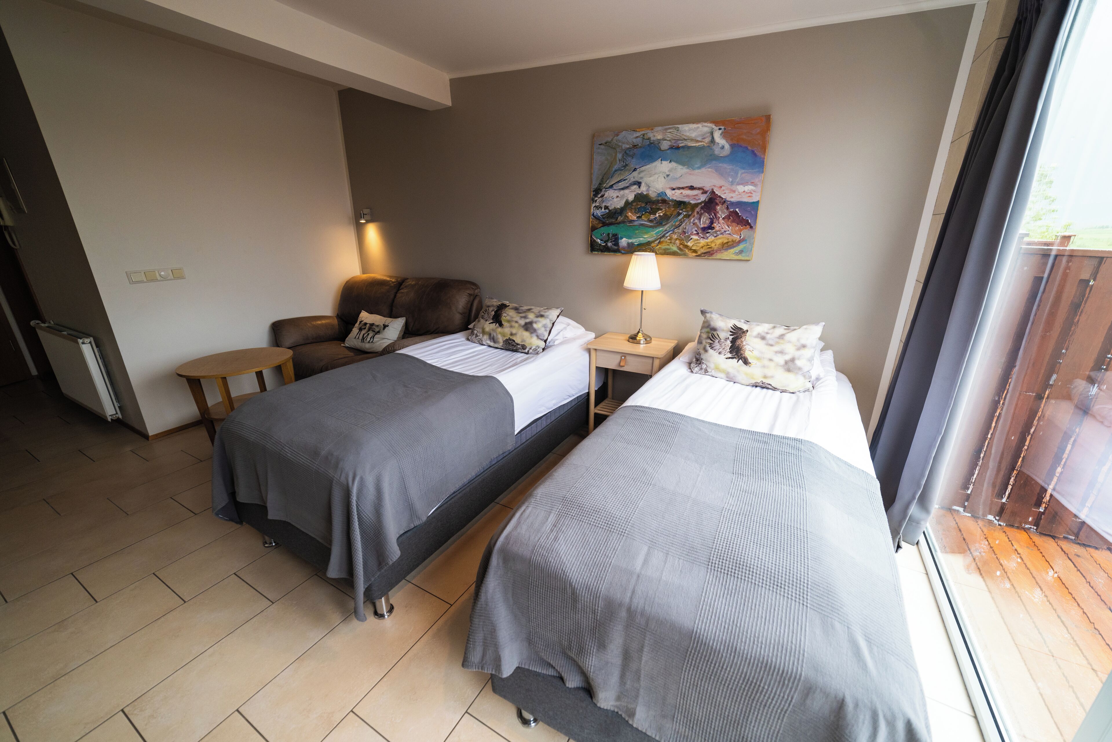 A hotel room with two beds, a painting on the wall, a lamp, and a view of the outdoors.