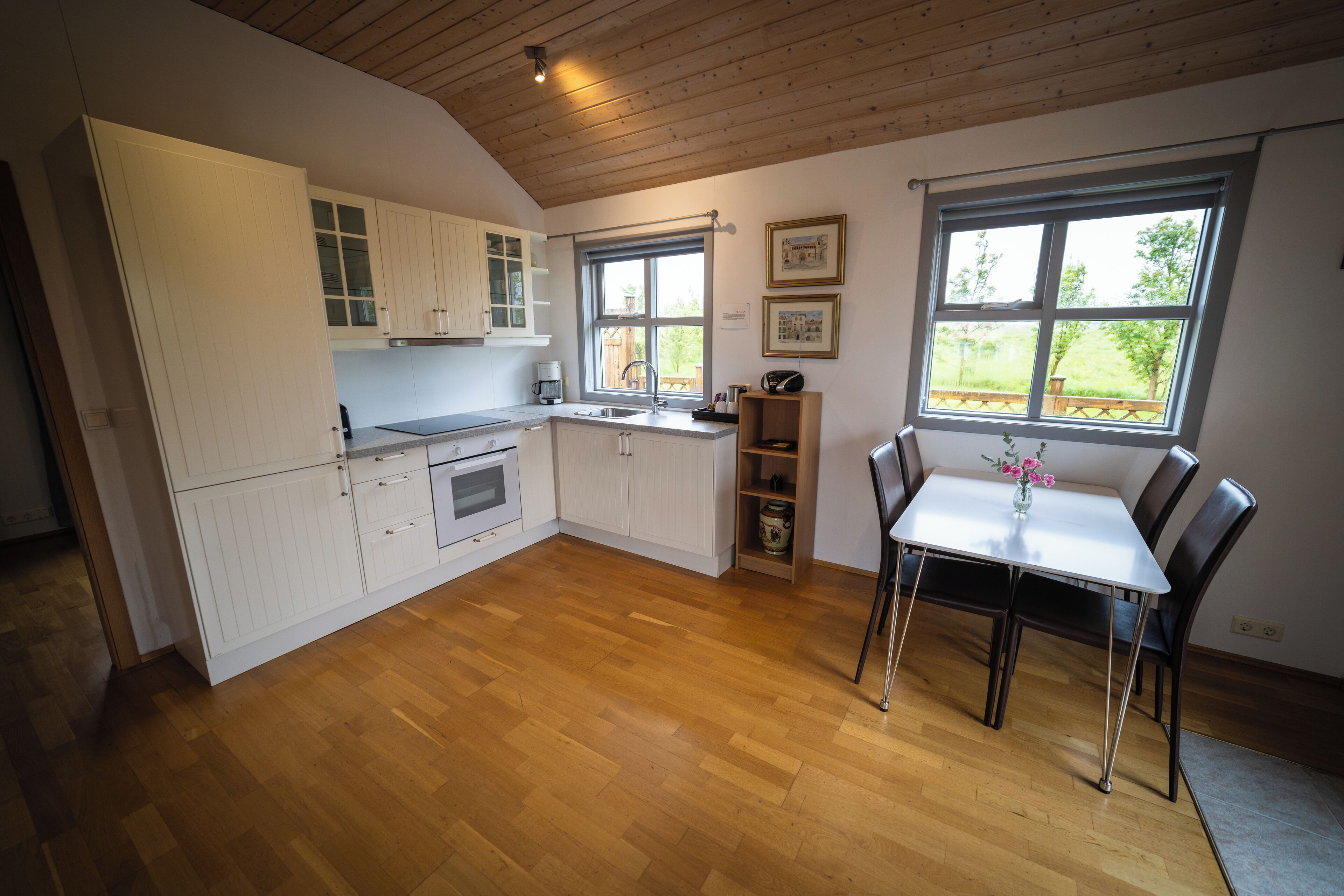A kitchen with wooden flooring, white cabinets, a dining table with chairs, and a window with a view of greenery.