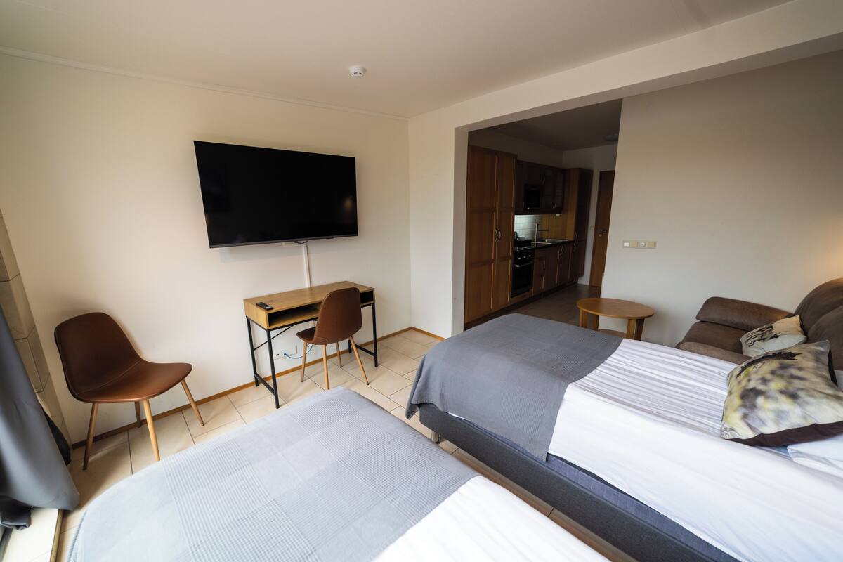 Ein Hotelzimmer mit zwei Betten, einem Schreibtisch mit Stuhl, einem Fernseher und einer Küchenzeile im Hintergrund.