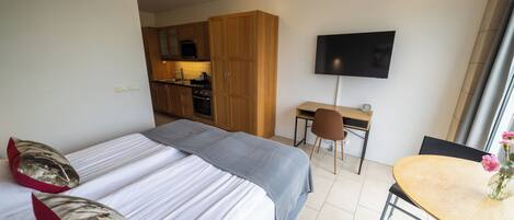 A hotel room with two beds, a desk, a chair, a TV, and a kitchen area.