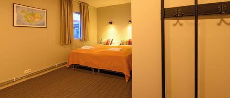 A hotel room with a bed, a window with curtains, a wall-mounted light, and a coat rack.