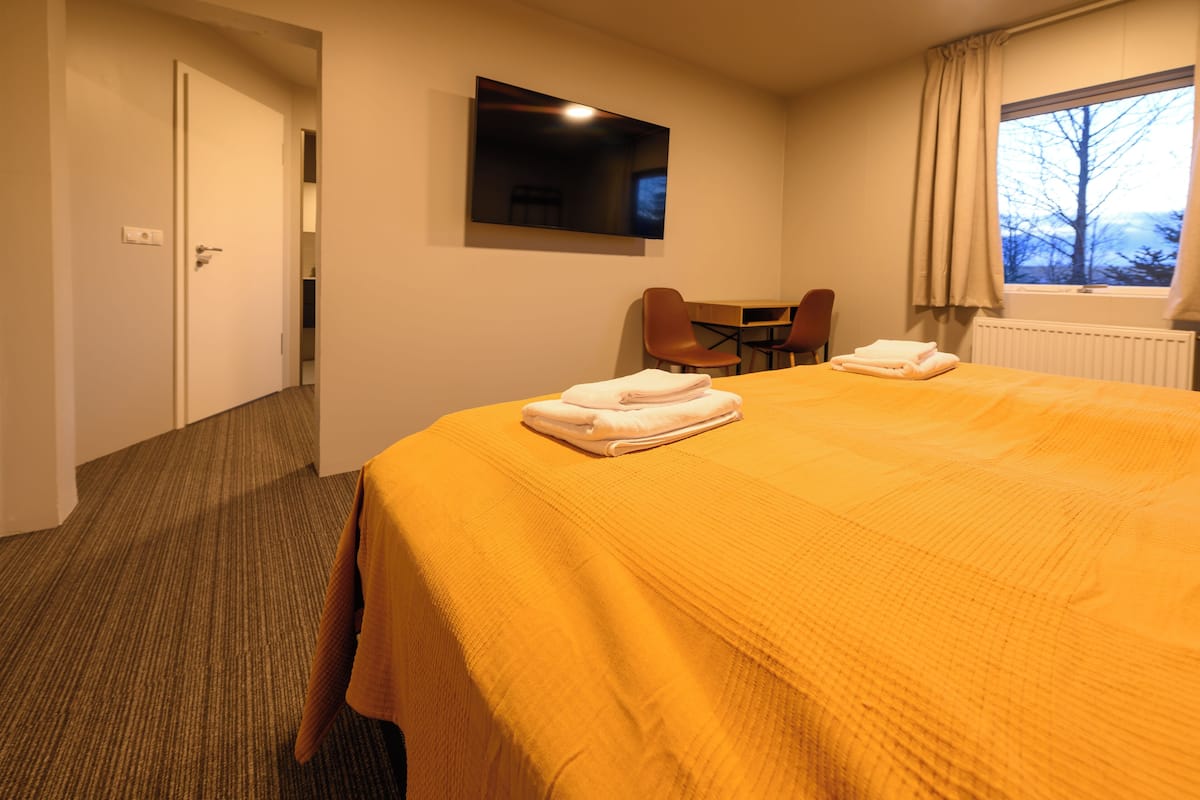 Ein Hotelzimmer mit einem großen Bett, einem flachbildfernseher an der Wand, einem Fenster mit Vorhängen und einem kleinen Tisch mit Stühlen.