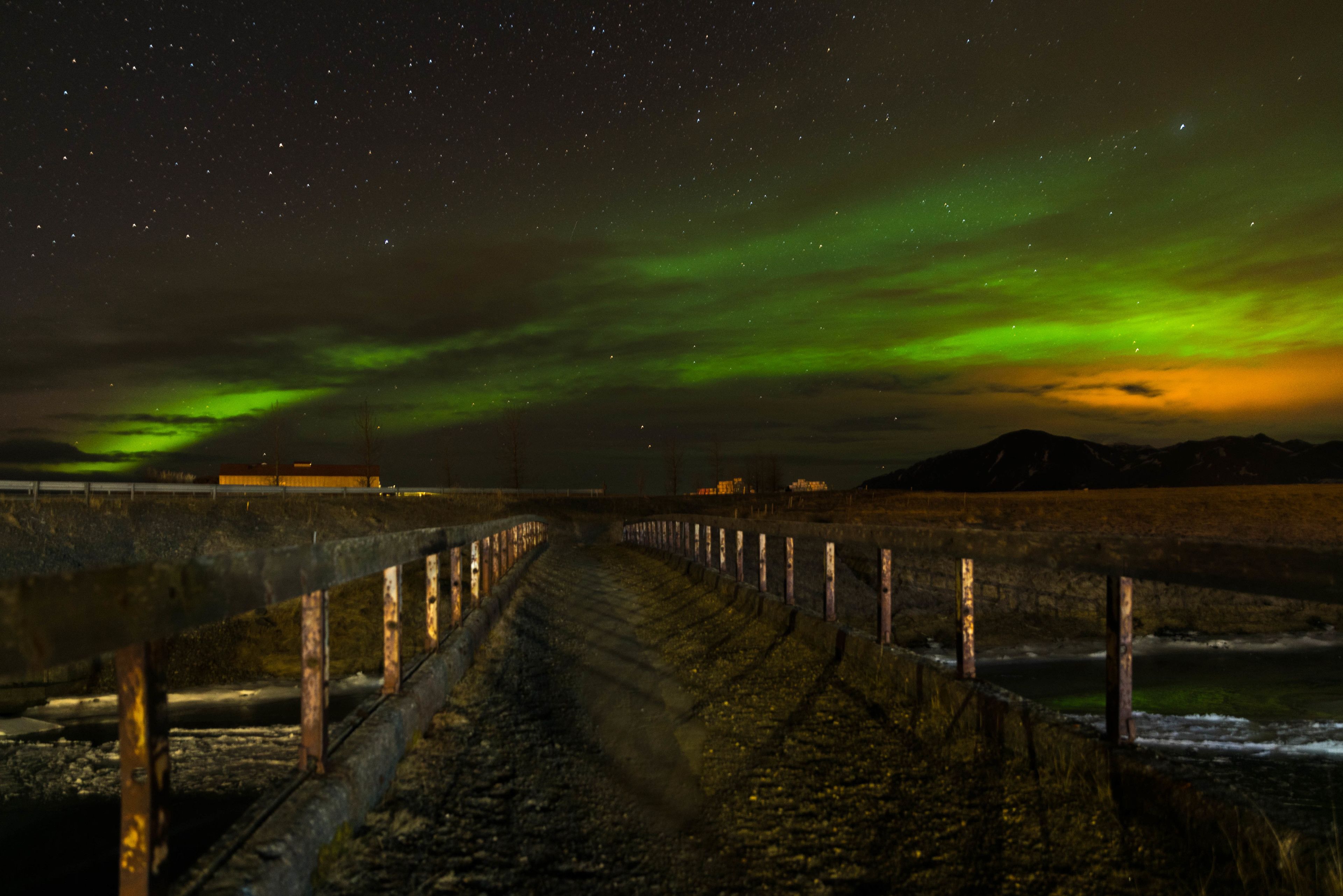 A nighttime view of a coastal area with a wooden walkway leading to the sea, under a starry sky with the aurora borealis.