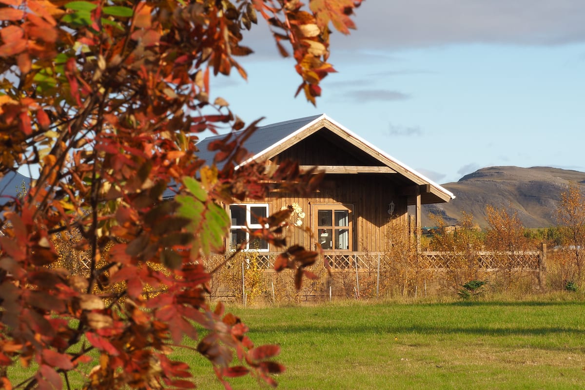 Eine Holzhütte mit Satteldach, umgeben von einem Holzzaun, vor einer Berglandschaft und blauem Himmel.