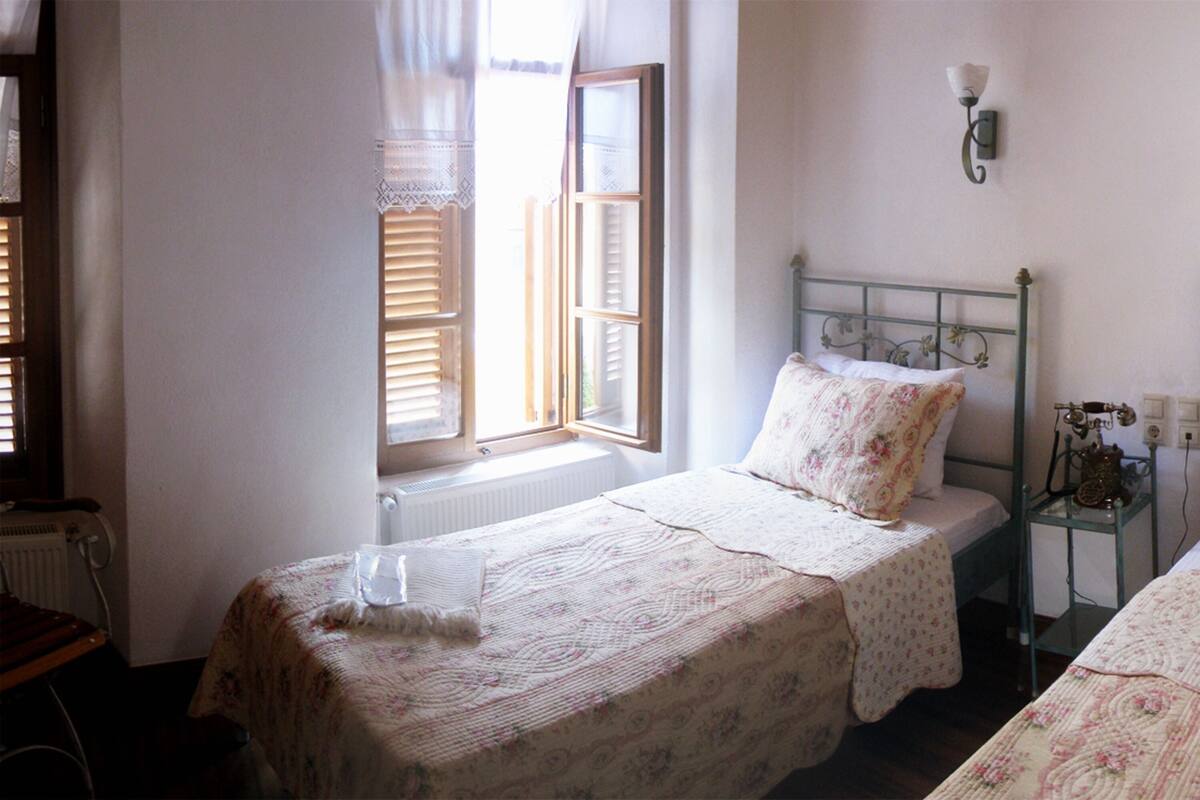 A room with two single beds, a small table with a lamp, and a window with blinds.