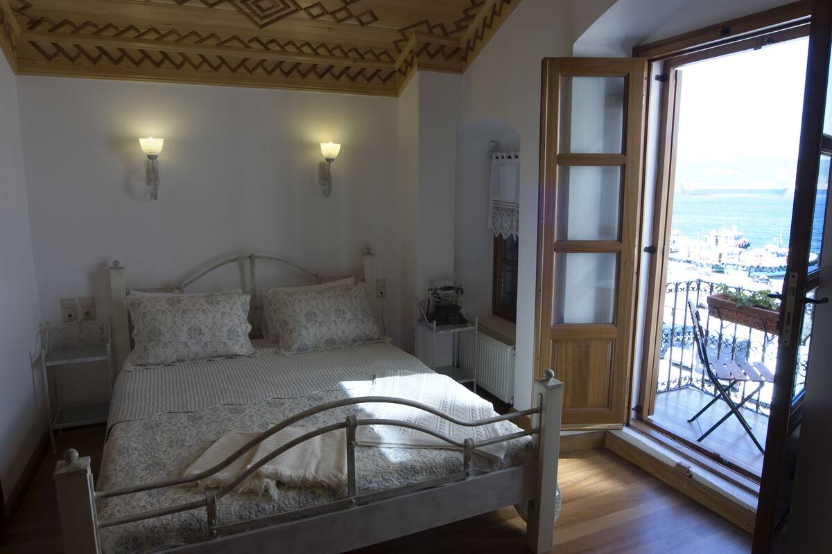 A bedroom with a bed, two wall-mounted lights, a radiator, and a view of the sea through an open door.