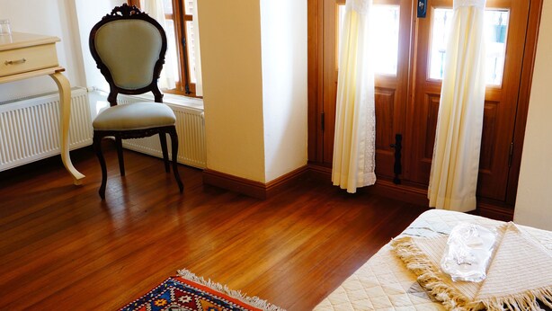 A room with a wooden floor, a bed with a quilt, a chair, a radiator, and a window with curtains. - Hotel Des Etrangers - Special Class (Çanakkale)
