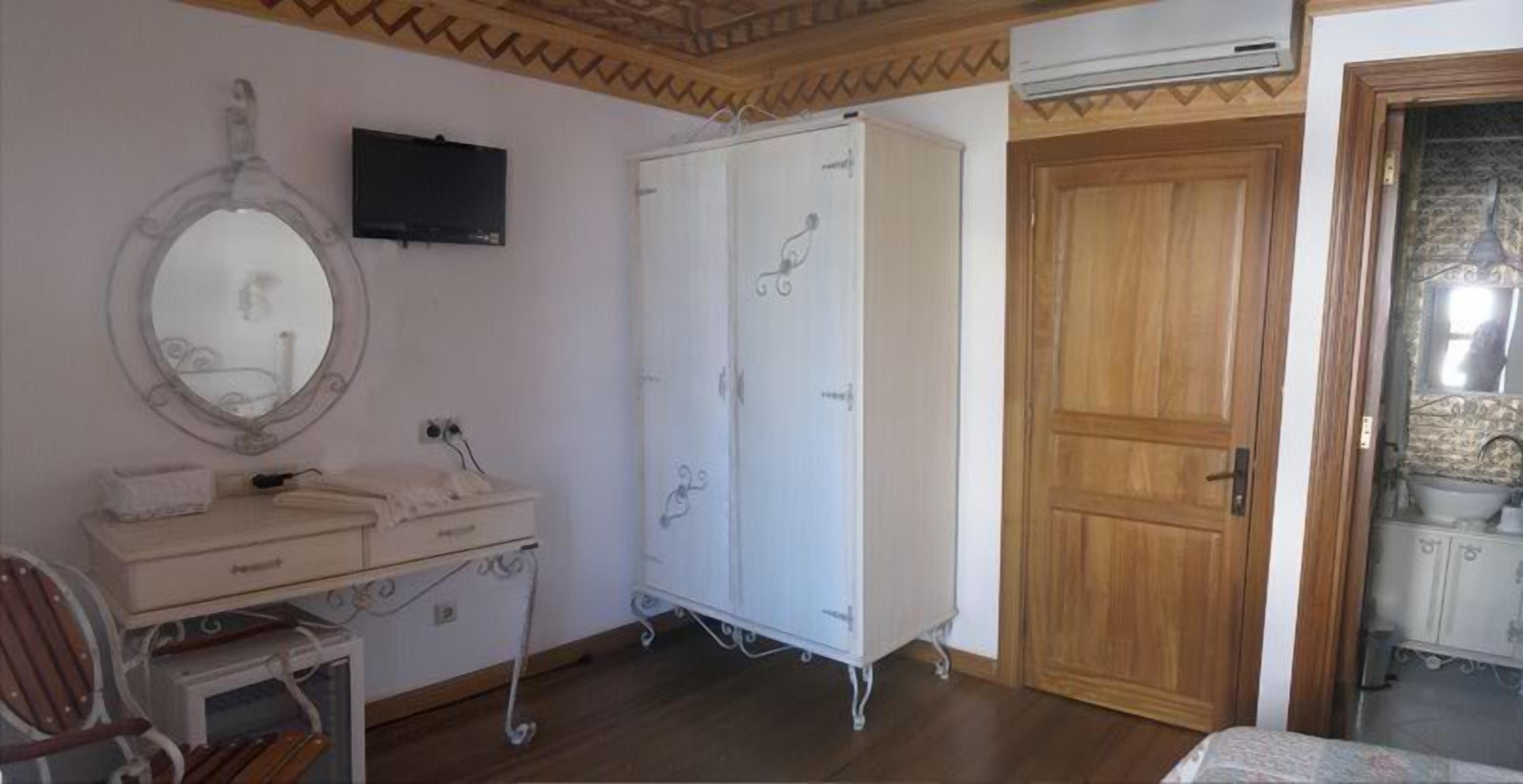 A room with a wooden door, a white wardrobe, a mirror, a small desk with a chair, and a television mounted on the wall.