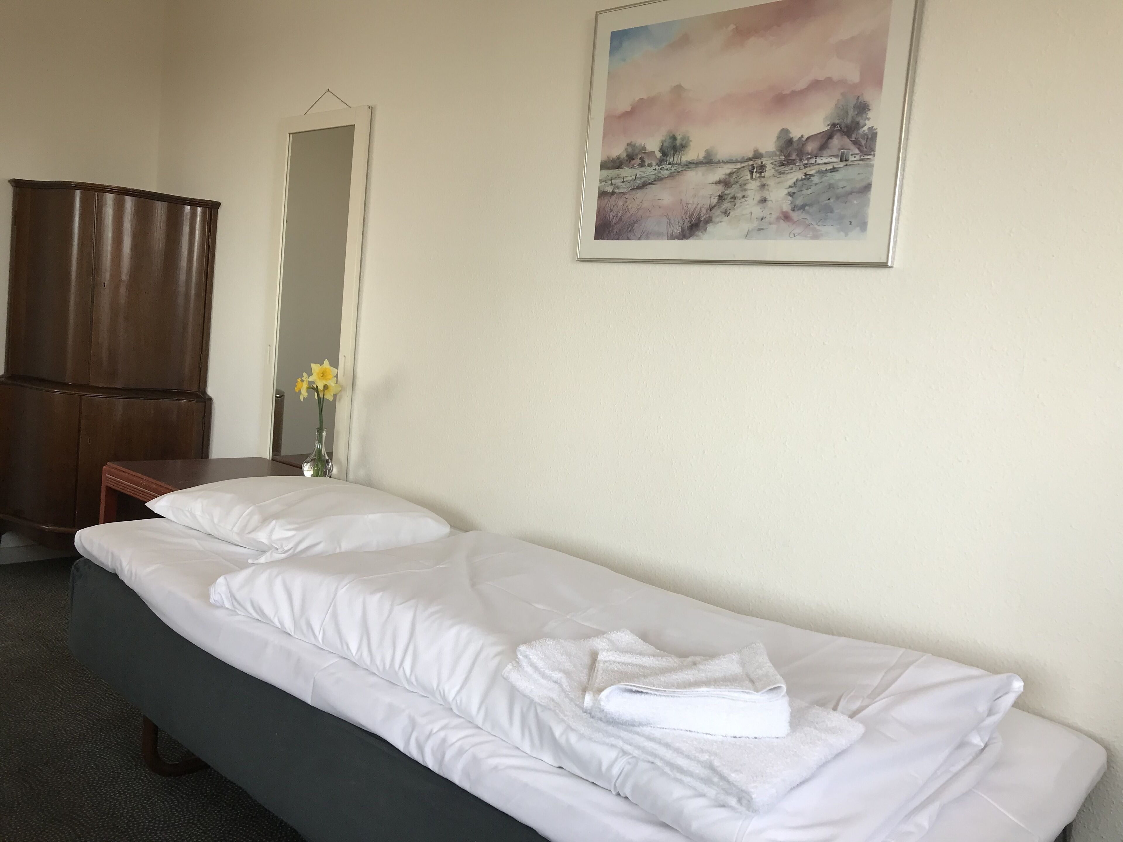 A hotel room with a single bed, a wooden wardrobe, a nightstand with a vase of flowers, and a landscape painting on the wall.