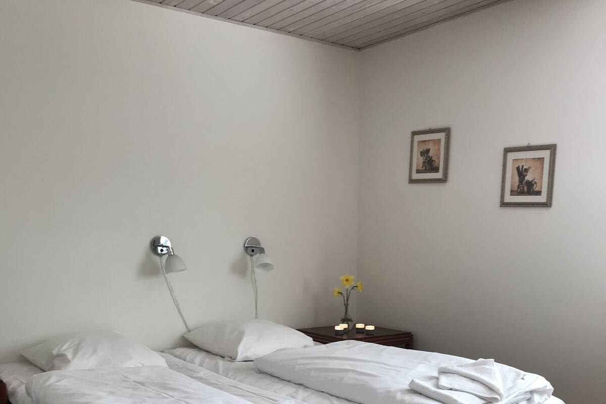 A double bed with white linens, a bedside table with a lamp, and two framed pictures on the wall.
