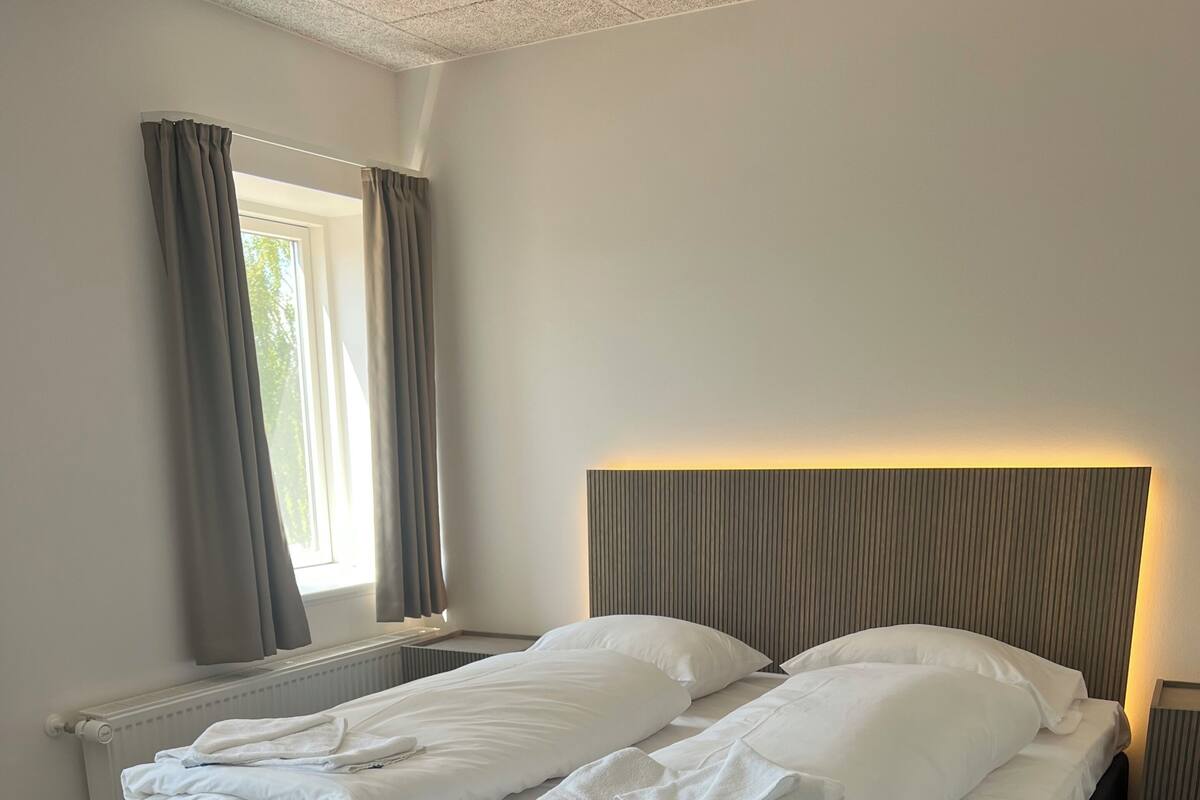 A double bed with a dark headboard and white bedding, a bedside table, a window with curtains, and a radiator.