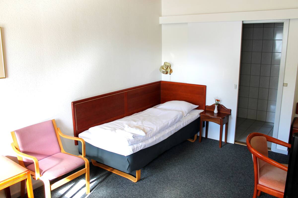 A single-bed room with a wooden headboard, a small wooden nightstand, a chair, and a desk.