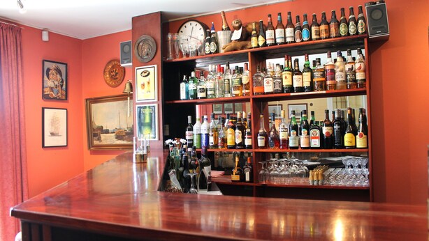 Un bar con estantes llenos de botellas de licor y vasos, un reloj y cuadros en la pared.