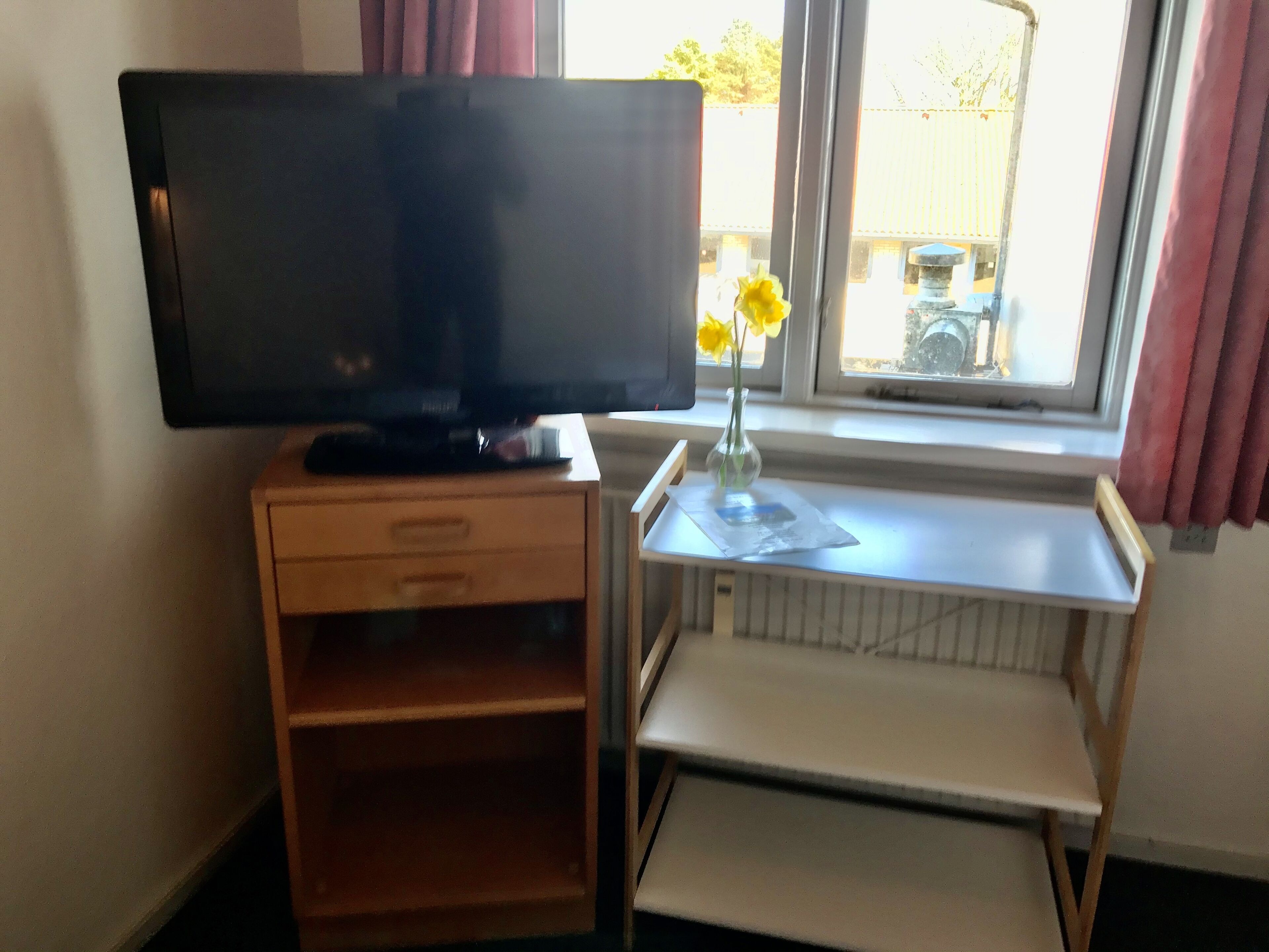 Un televisor sobre un mueble de madera, una mesita con un florero y una ventana con cortinas.