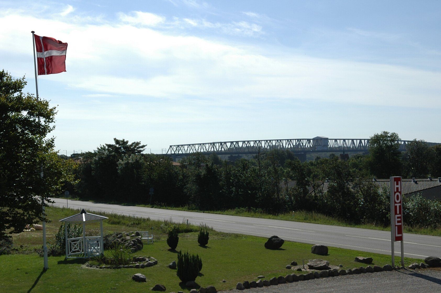 A hotel sign with a red and white design, a flagpole with a flag, a road, and a bridge in the background.