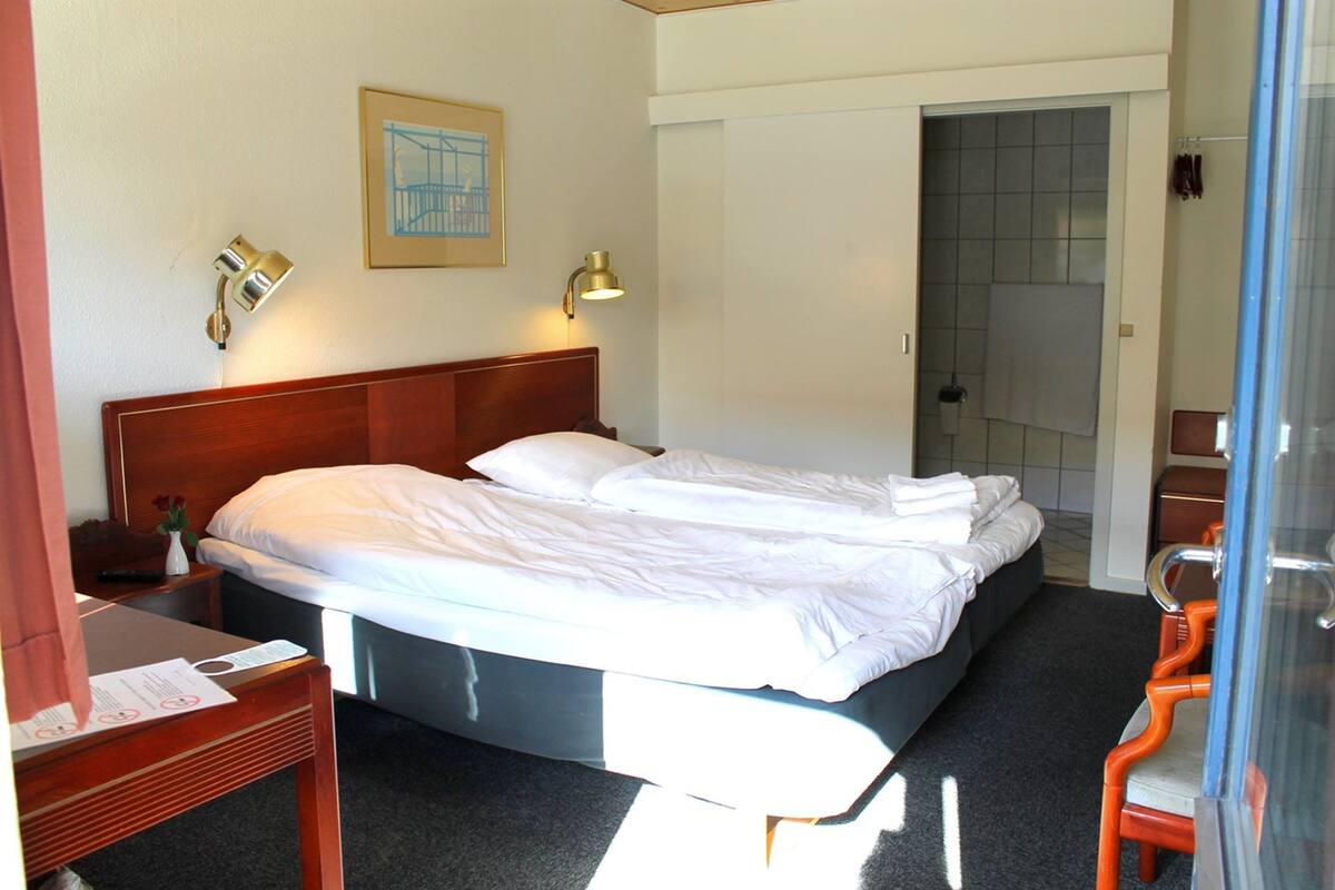 A hotel room with a double bed, a wooden headboard, two bedside lamps, a framed picture on the wall, and a shower area visible through an open door.