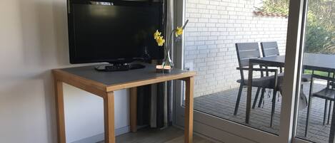 A modern living room with a television on a wooden table, a vase with yellow flowers, and a view of an outdoor patio with a brick wall and a table and chairs.