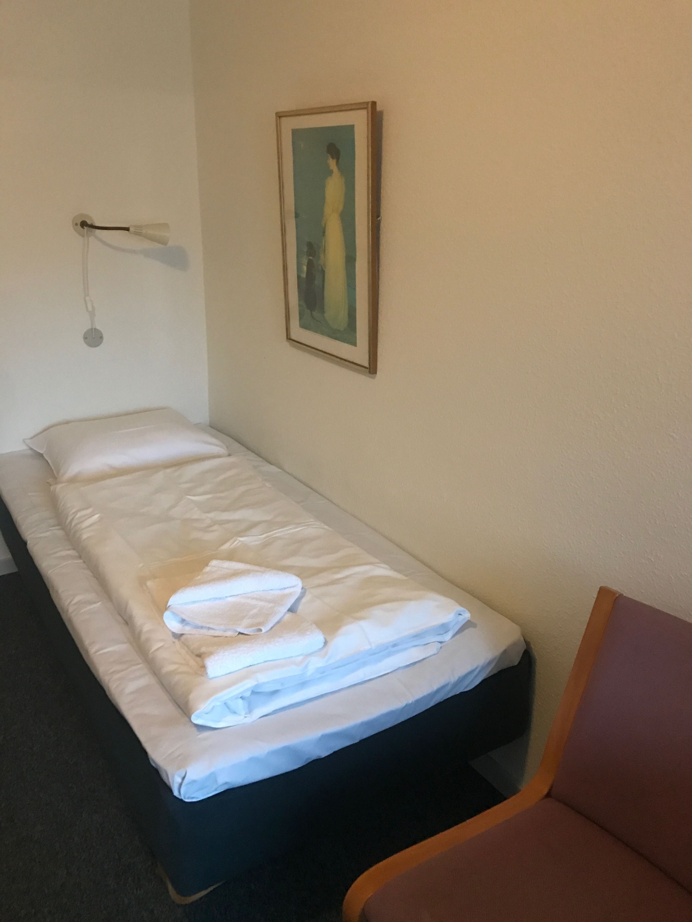 Una cama individual con ropa de cama blanca y una toalla doblada, una lámpara de noche y un cuadro en la pared.