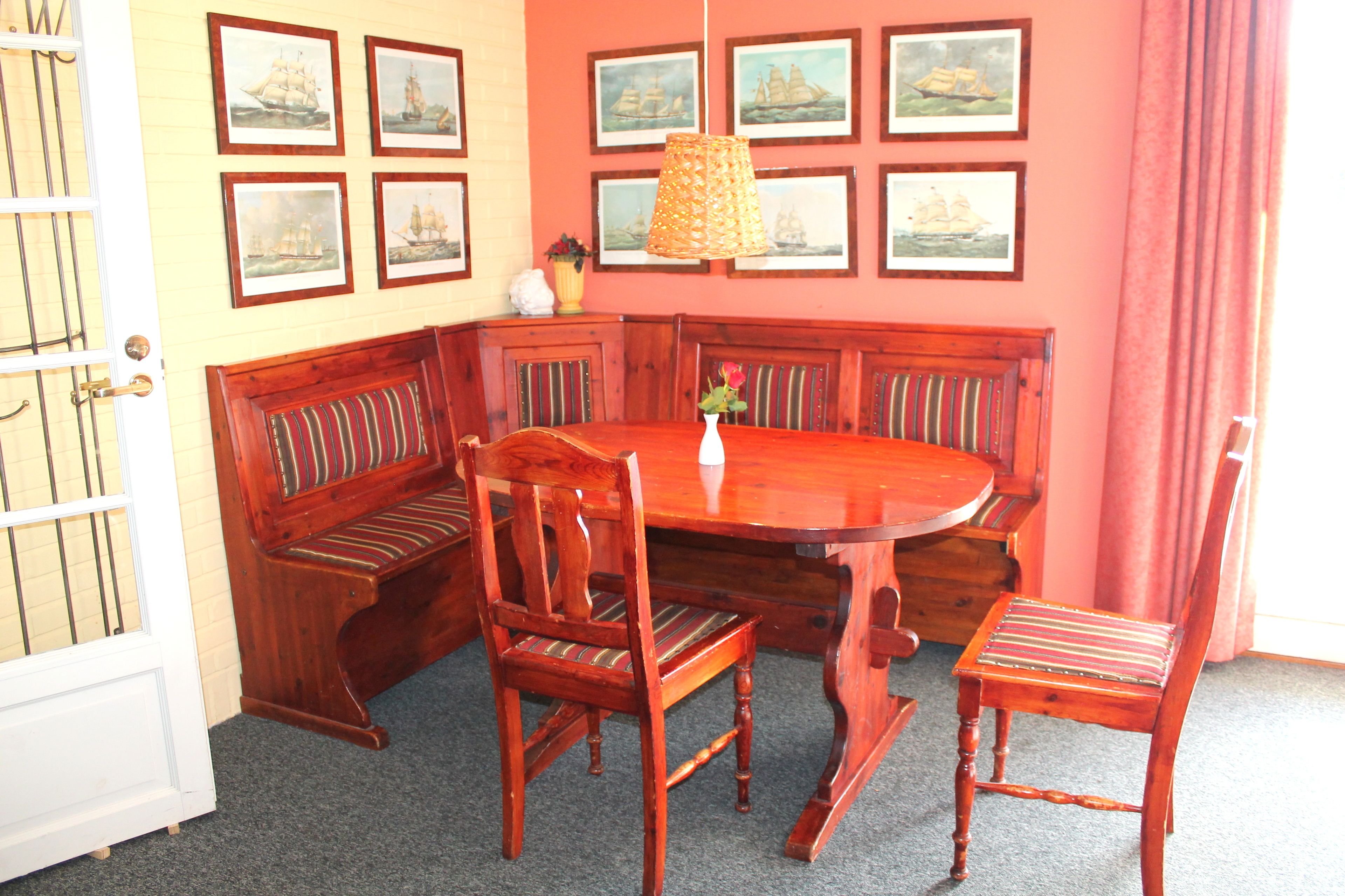 A dining area with a wooden bench seating around a central table, framed maritime paintings on the wall, and a hanging lamp.