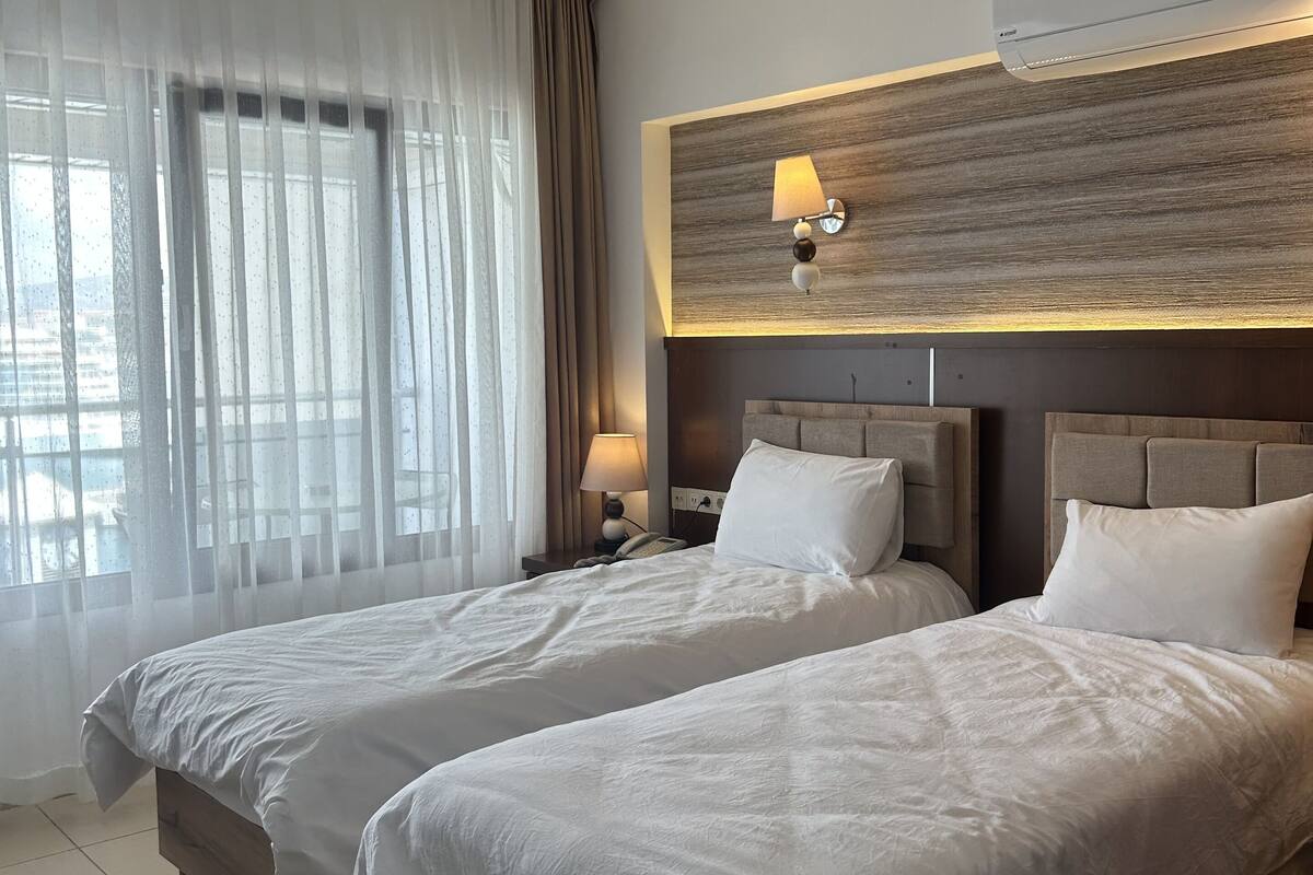A hotel room with two beds, a large window with sheer curtains, a wall-mounted lamp, and an air conditioning unit.