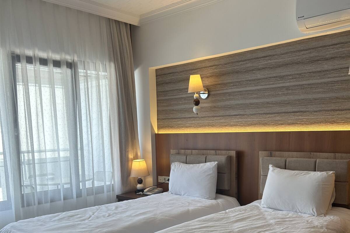 A hotel room with two beds, a wooden headboard, a wall-mounted lamp, and a window with sheer curtains.