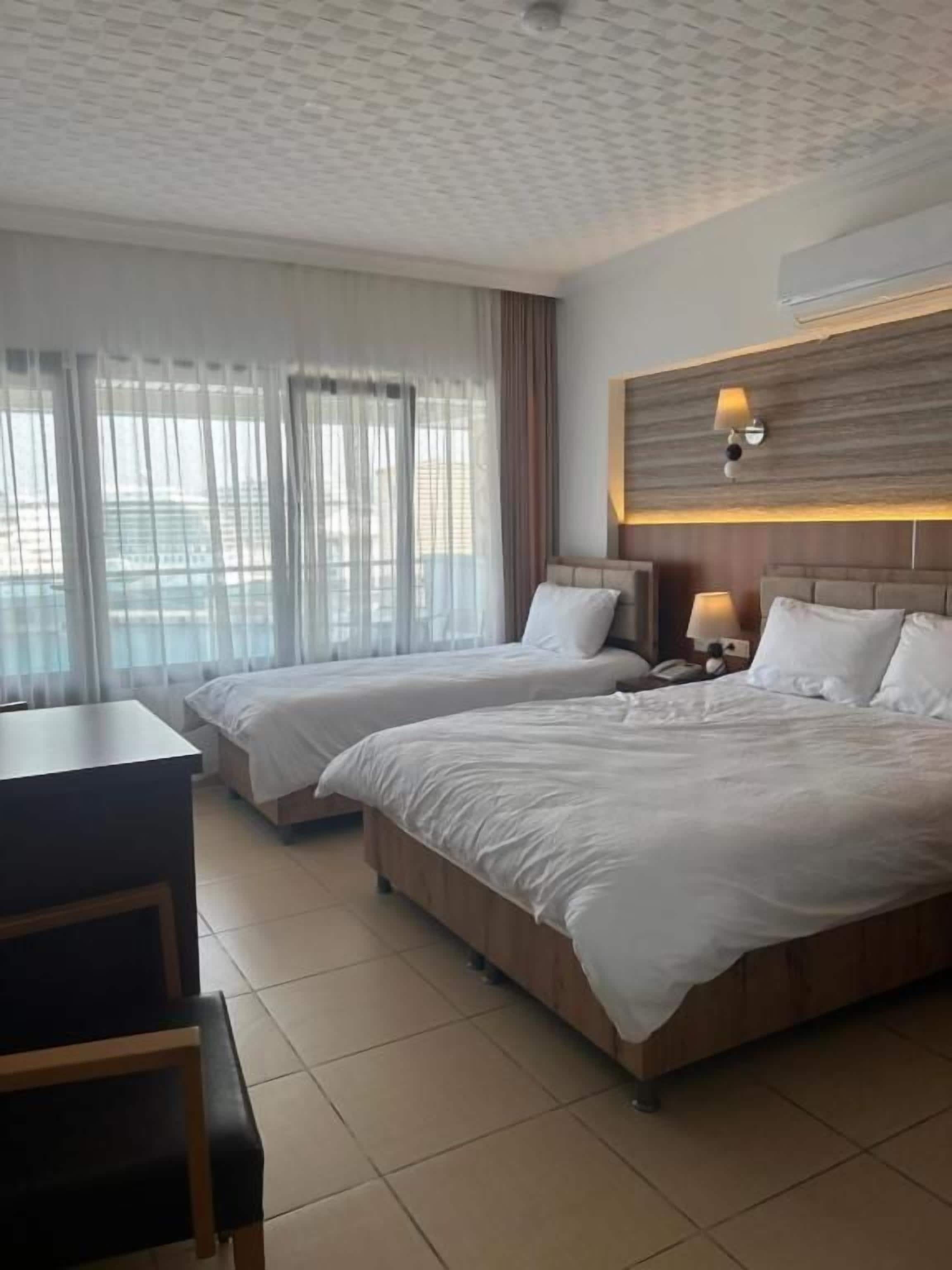 Habitación de hotel con dos camas, un escritorio y vistas de la ciudad a través de amplias ventanas.