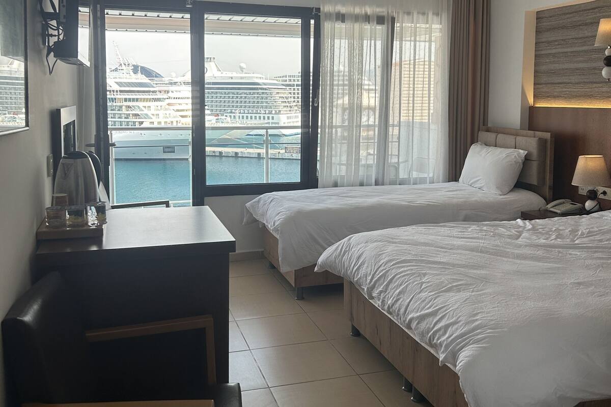 A hotel room with two beds, a desk, a chair, a lamp, and a view of a cruise ship and water.