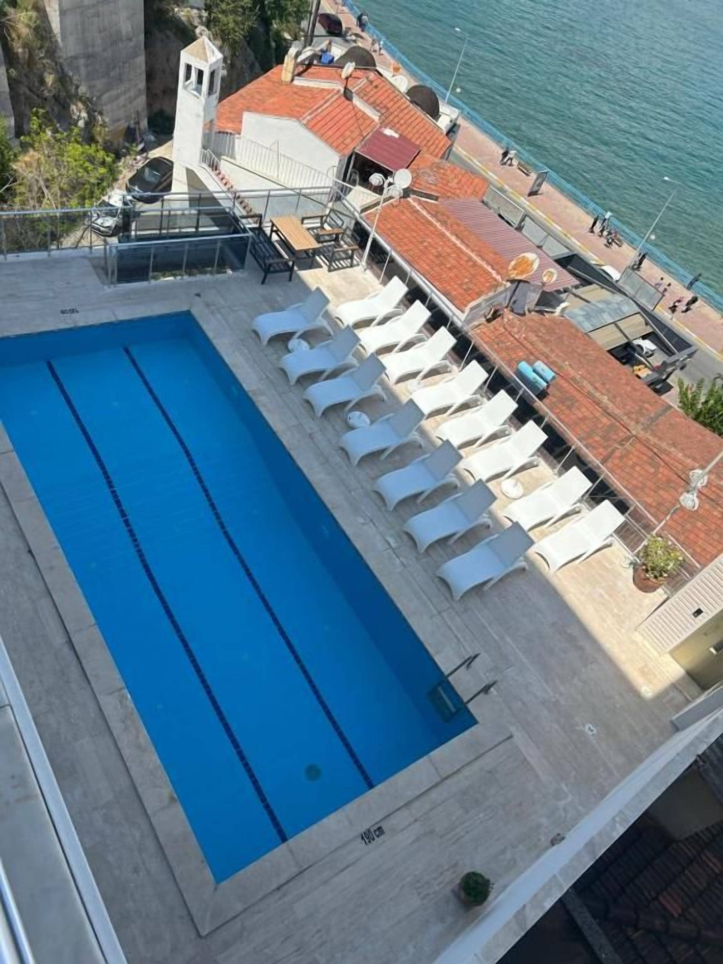 A rooftop swimming pool with lounge chairs, a view of the sea, and a building with a red-tiled roof.