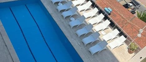Una piscina en la azotea con sillones de descanso, vista al mar y un edificio con techo de tejas rojas.