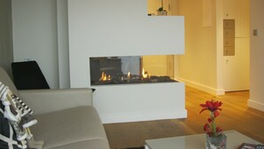 Una sala de estar moderna con chimenea, un sofá, una mesa de centro con un jarrón de flores y una zona de cocina al fondo.
