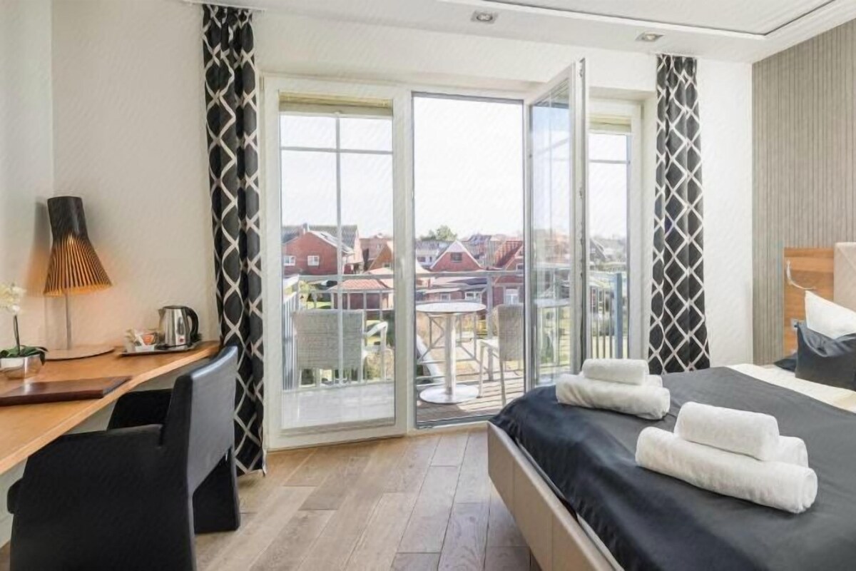 Habitación de hotel con cama, escritorio, silla, lámpara y vistas a través de un ventanal.