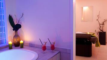 A spa room with a bathtub, two candles, a vase with a plant, and two drinks on the side.