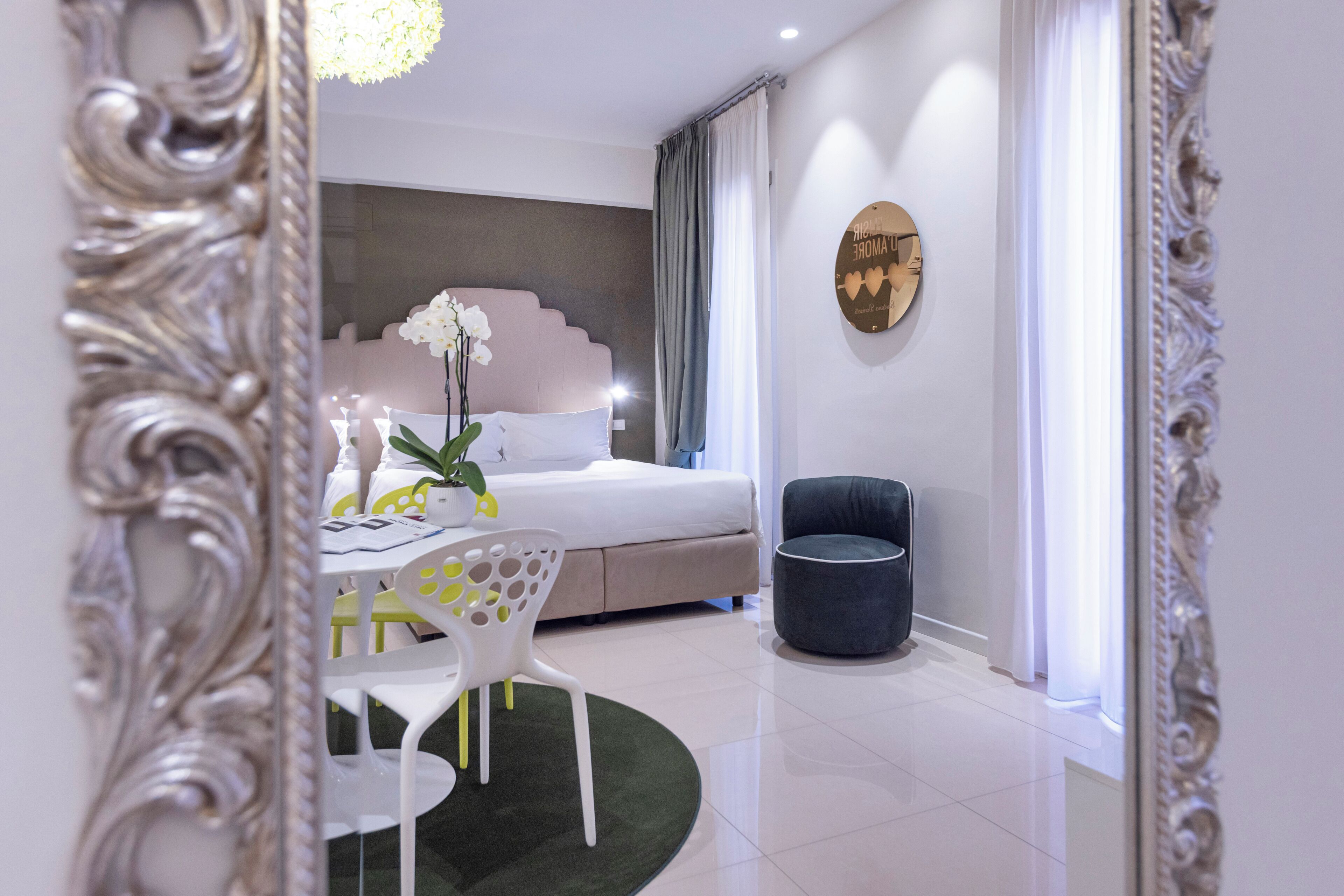 A modern hotel room with a bed, a dining table with chairs, a potted plant, and a decorative wall piece.