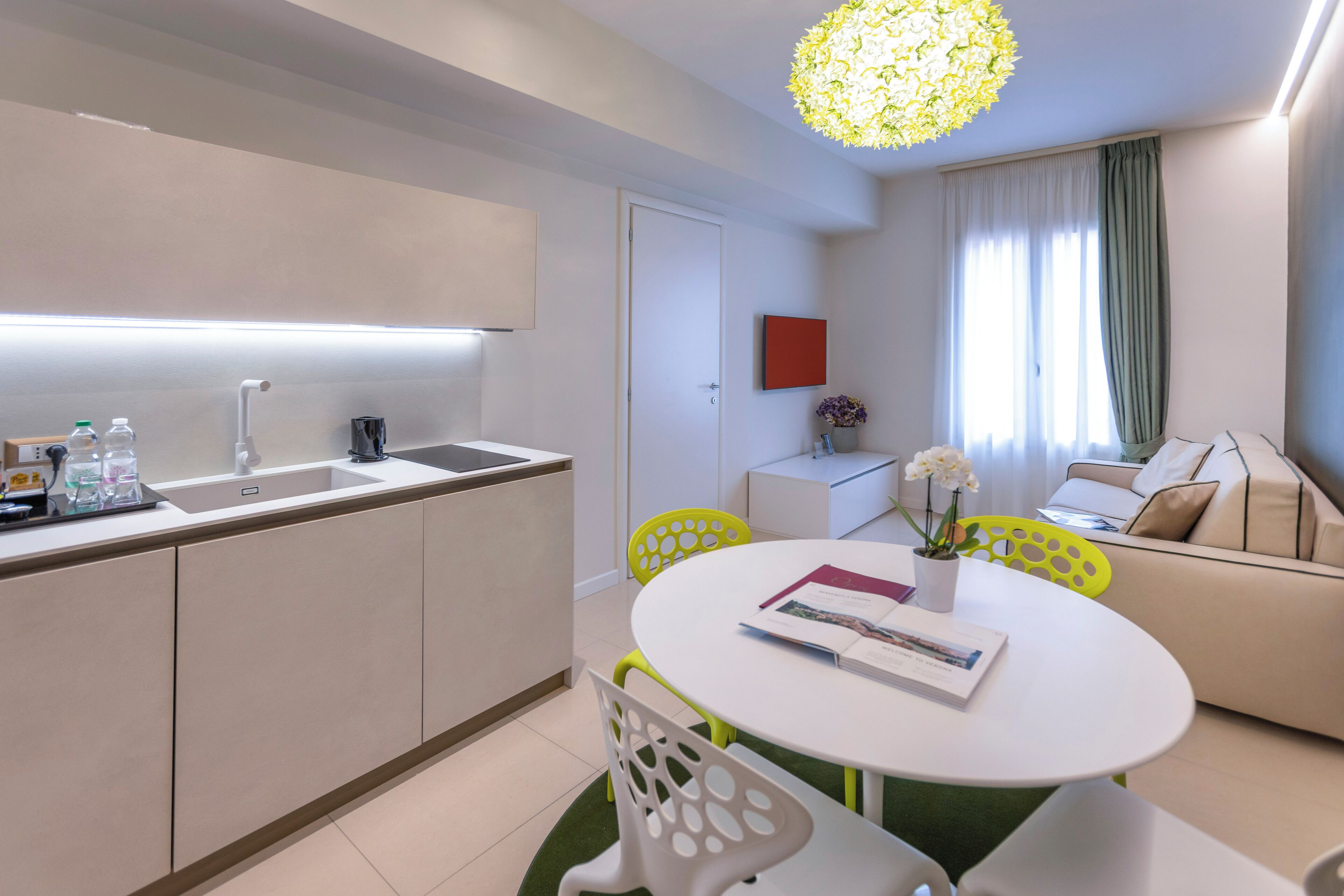 A modern kitchen with a white countertop and sink, a dining area with a white table and yellow chairs, and a living area with a beige sofa and a green curtain.