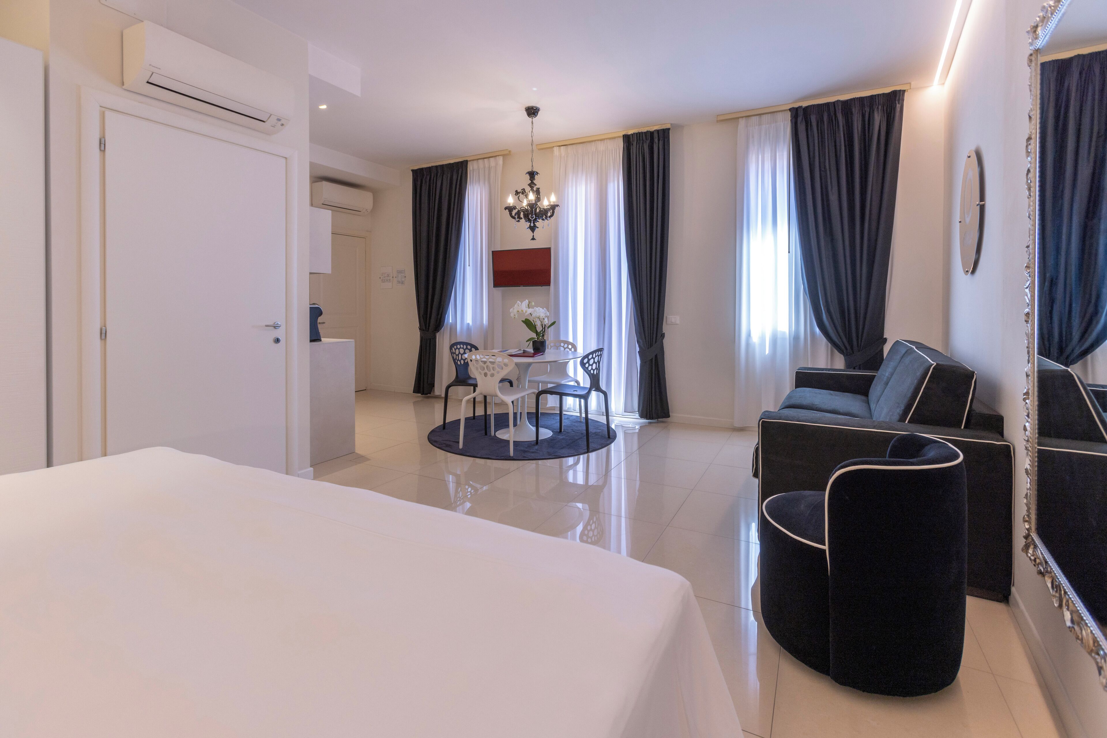 A hotel room with a bed, a dining table with chairs, a sofa, a mirror, and a chandelier.