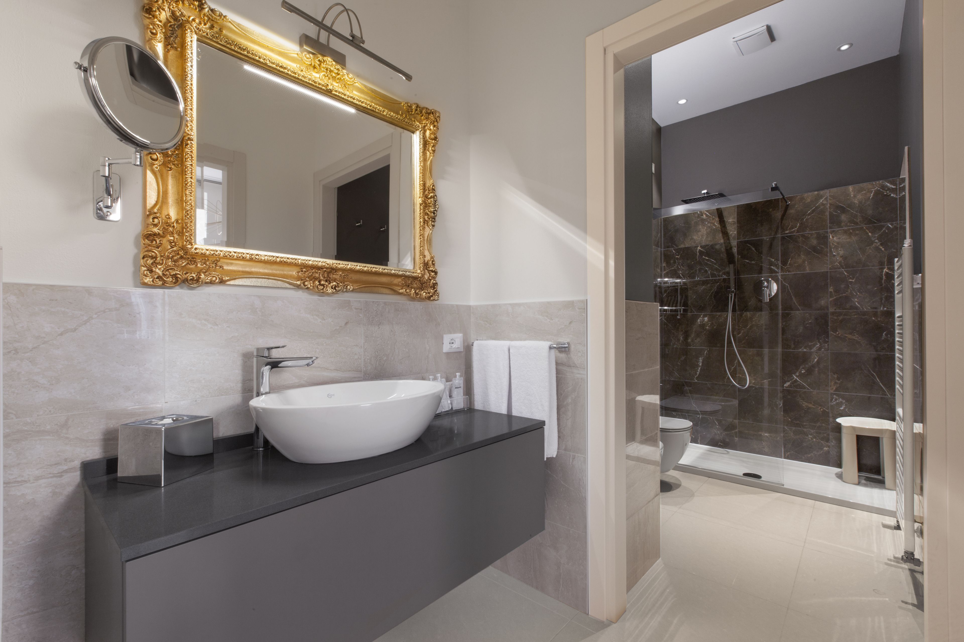 Un baño moderno con un lavamanos ovalado grande, un espejo con marco dorado y una ducha a ras de suelo con baldosas de mármol.